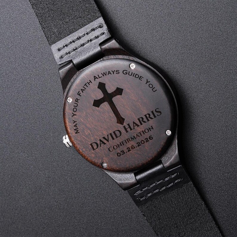 May include: A wooden wristwatch with a dark brown face and black leather strap. The watch face features a cross design and the engraved text: "MAY YOUR FAITH ALWAYS GUIDE YOU" and "DAVID HARRIS CONFIRMATION 03.26.2026".
