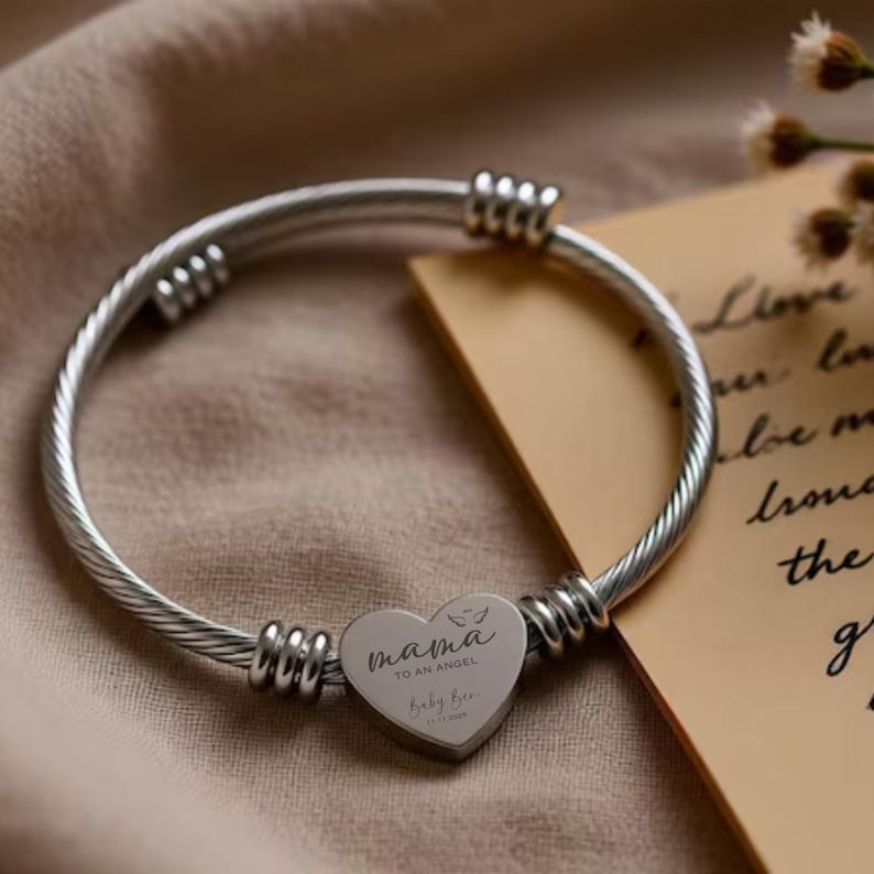 May include: Silver-toned bangle bracelet with a heart-shaped charm. The charm is engraved with the words "mama TO AN ANGEL" and "Baby Bee" along with a date. The bracelet has a twisted cable design.