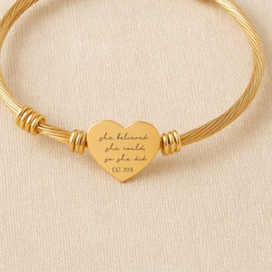 May include: Gold-coloured bangle bracelet featuring a heart-shaped charm. The charm is engraved with the words "she believed she could so she did" and "EST 2008". The bracelet has a twisted rope design.