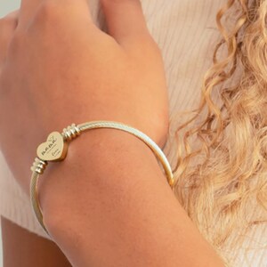 May include: Gold-toned bangle bracelet with a heart-shaped charm. The charm is engraved with the word "Mama" and other script. The bracelet has a cable-style design and is worn on an arm.