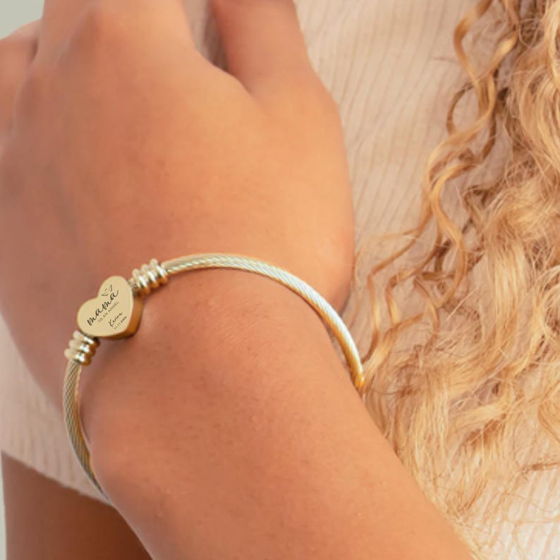 May include: Gold-toned bangle bracelet with a heart-shaped charm. The charm is engraved with the word "Mama" and other script. The bracelet has a cable-style design and is worn on an arm.