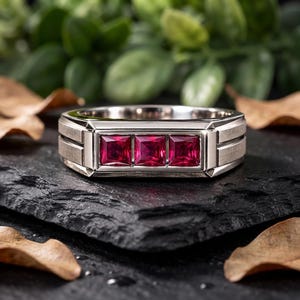 Puede incluir: Un anillo de tono plateado con tres rubíes rojos vibrantes de corte cuadrado, dispuestos horizontalmente en fila. La banda tiene un acabado de metal cepillado con ranuras horizontales distintivas en los lados, creando un aspecto estructurado.