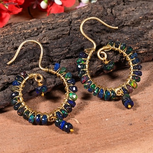 Peut inclure: Une paire de boucles d'oreilles créoles dorées. Chaque boucle d'oreille présente un design circulaire avec de petites pierres précieuses facettées bleu foncé et vertes. Les pierres sont enroulées de fil d'or, et les boucles ont une fermeture à crochet.