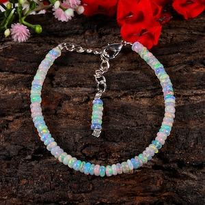 Puede incluir: Una pulsera de cuentas de ópalo con una cadena y un cierre de plata. La pulsera presenta pequeñas cuentas redondas de ópalo en tonos blancos, azules y verdes. La pulsera se exhibe sobre una superficie oscura y texturizada con detalles florales.