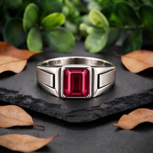 May include: A silver ring featuring a rectangular, emerald-cut red gemstone set in the center. The band has a smooth, polished finish with a distinct geometric design on the sides. The interior of the band is clearly stamped with the text 925.