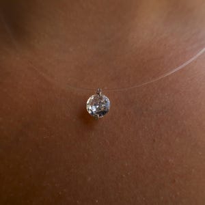 Puede incluir: Un colgante de piedra preciosa redonda y facetada, transparente, suspendido de un collar de hilo de nailon fino. La piedra presenta un corte brillante con múltiples superficies reflectantes que captan la luz, sujeta por un engaste metálico sencillo.