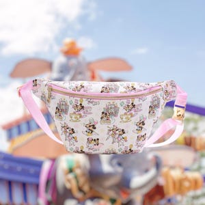 May include: A white fanny pack featuring a repeating pattern of Mickey and Minnie Mouse characters, castles, and teacups. The bag has a light pink adjustable strap with a gold-tone buckle and a gold-tone zipper closure across the top front.