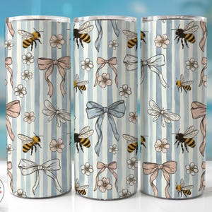 May include: Three stainless steel tumblers with a repeating pattern of bees, bows, flowers, and dragonflies on a light blue striped background. The design includes pink, light blue, and yellow accents. The tumblers are cylindrical and have a shiny finish.