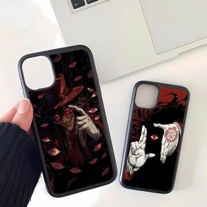 May include: Black phone cases featuring anime-style illustrations. One case displays a character in a red hat and sunglasses surrounded by red eyes. The other case shows a hand gesture with a red eye and a symbol.