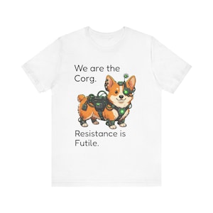 May include: A white short-sleeved t-shirt featuring a graphic of a cyborg Corgi dog wearing mechanical gear and a glowing green eye piece. The text printed on the shirt reads, We are the Corg. Resistance is Futile. The design is centered on the chest.