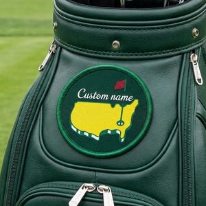 Op de afbeelding: Een donkergroene golftas met zilveren ritsen en een ronde patch met een gele kaart en een rode vlag. De tekst "Custom name" staat boven de kaart. De tas heeft het PGA-logo.