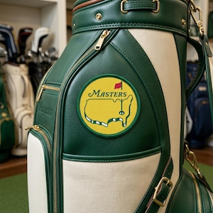Puede incluir: Bolsa de golf verde y crema con cremalleras y herrajes dorados. La bolsa presenta un parche con el logotipo de Masters y la palabra "Augusta". Tiene una parte superior de cuero marrón y una sección de lona crema.