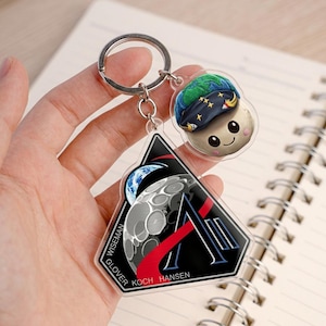 May include: A clear acrylic keychain with two charms. One charm features a cartoon mushroom character wearing a blue hat with stars. The other charm is a black and red triangle with a moon and Earth design, and the names GLOVER, WISEMAN, KOCH, and HANSEN.