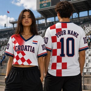 A woman wearing a white and red Croatia soccer jersey stands in front of a stadium, with a man wearing a similar jersey visible behind her.