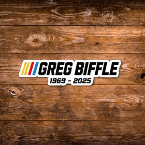 May include: A white sticker featuring the name "GREG BIFFLE" in bold black text, with a multi-coloured stripe design above. The text "1969 - 2025" is below the name. The sticker is placed on a wooden surface.