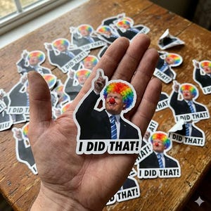 May include: A sticker featuring a caricature of a man in a suit with a rainbow clown wig and a red nose, pointing upwards. The text "I DID THAT!" is below the image. Multiple stickers are scattered on a wooden surface.