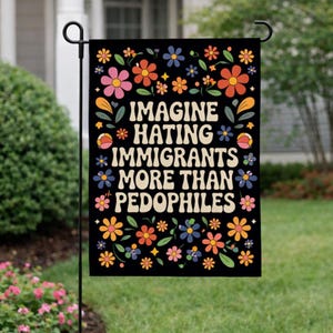 Op de afbeelding: Een zwarte tuin vlag met de tekst "IMAGINE HATING IMMIGRANTS MORE THAN PEDOPHILES" in het wit, omringd door kleurrijke bloemen. De vlag hangt aan een zwarte metalen standaard in een tuin.