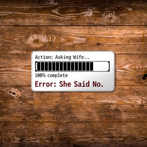 May include: A rectangular white sticker with black text and a loading bar graphic. The text reads "Action: Asking Wife..." and "100% complete." Below, it says "Error: She Said No." The sticker is set against a wooden background.