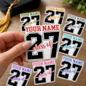 May include: A collection of graduation-themed stickers with the text "YOUR NAME" and "class of 27". The stickers feature a black and white design with various accent colors, including red, green, and blue. A hand holds one sticker.