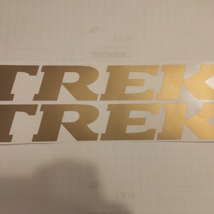 May include: Two gold-colored "TREK" decals are displayed on a white background. The decals are in a bold, sans-serif font, with the letters appearing slightly shadowed. The decals are likely for a bicycle or outdoor equipment.