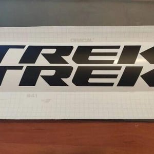Könnte beinhalten: Zwei schwarze "TREK" Aufkleber, einer über dem anderen, werden auf weißem Hintergrund angezeigt. Das Wort "TREK" ist in einer fetten, serifenlosen Schriftart gehalten. Der Hintergrund hat ein Rastermuster und das Wort "ORACAL" ist sichtbar.