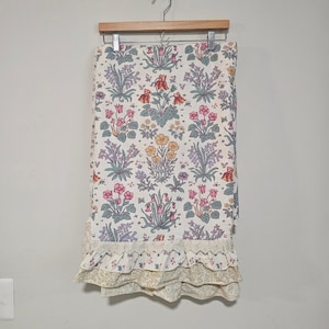 Peut inclure: Un panneau de tissu rectangulaire présentant un motif floral répétitif avec des fleurs roses, jaunes et violettes sur fond blanc. Le bord inférieur est fini avec une bordure en dentelle crème et deux volants superposés à motifs floraux.