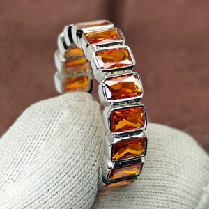 May include: A silver ring featuring a series of rectangular, orange-colored gemstones. The gemstones are set in a row, creating a continuous band of color. The ring is held against a neutral background, highlighting the gemstones' brilliance.