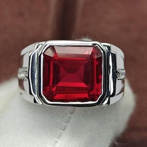 May include: A silver-toned ring featuring a large, rectangular, faceted red gemstone set in the center. The band has a polished metallic finish with small, clear, sparkling stones embedded along the sides of the shank, creating a detailed design.