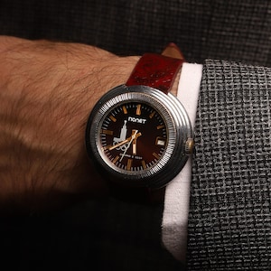 Puede incluir: Un reloj de pulsera vintage con caja plateada y esfera marrón oscuro. El reloj tiene agujas y marcadores dorados, y una correa de cuero roja. La esfera del reloj tiene la palabra "ПОЛЕТ" y un logotipo. El reloj está en una muñeca.