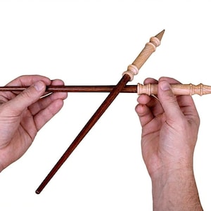 以下が含まれることがあります： 白い背景に2本の木製の杖が写っています。1本は濃い茶色でまっすぐ、もう1本は明るい茶色で装飾的な柄と尖った先端が付いています。杖は交差しており、対照的な色とデザインが際立っています。