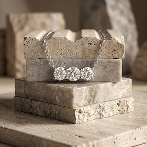 May include: A silver necklace featuring three round, brilliant-cut diamonds. The necklace is displayed on a stack of textured, light-colored stone blocks. The diamonds sparkle, reflecting light.