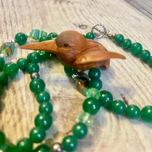 May include: A handcrafted necklace featuring a carved wooden bird pendant. The necklace has strands of vibrant green beads and accents of colorful glass beads. The bird pendant is a warm brown color, and the necklace has a silver clasp.