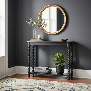 Może przedstawiać: Czarna konsola z toczonymi nogami, dolną półką i okrągłym lustrem w złotej ramie. Stół jest ozdobiony rośliną, książkami i małą miseczką. Na podłodze leży dywan z geometrycznym wzorem.