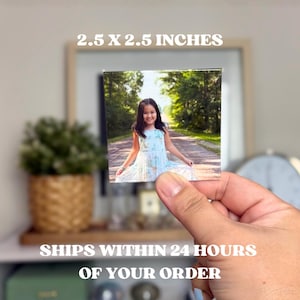 May include: A square photograph, 2.5 x 2.5 inches, featuring a young girl in a light blue dress, held by a hand. The image within the photo shows the girl smiling on a path lined with trees. The text "2.5 X 2.5 INCHES" is at the top.