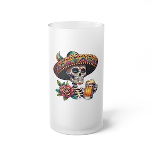 May include: A white frosted glass features a colorful Day of the Dead skeleton wearing a sombrero and holding a beer stein. A red and green rose is next to the skeleton. The design is a vibrant illustration.