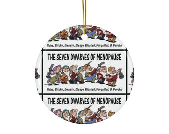 May include: A round, white ceramic ornament with a gold string for hanging. The ornament features an illustration of the Seven Dwarves with the text "THE SEVEN DWARVES OF MENOPAUSE" and the phrases "Itchy, Bitchy, Sweaty, Sleepy, Bloated, Forgetful, & Psycho".