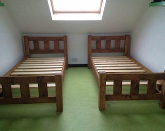 May include: Two wooden twin beds with a natural finish are positioned side-by-side on a green floor. Each bed has a headboard and footboard with rectangular cutouts. The beds are in a room with a skylight.