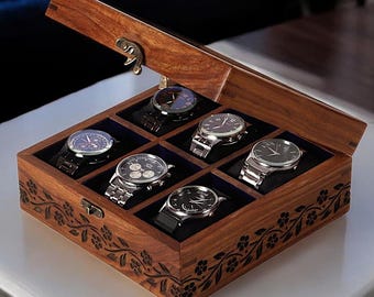 Puede incluir: Una caja de reloj de madera con tapa con bisagras, que contiene seis relojes de pulsera. La caja es marrón con tallas florales y un interior azul oscuro. Los relojes tienen esferas y correas plateadas y negras.