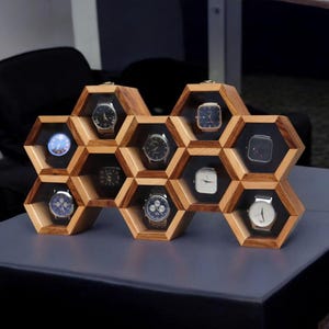 May include: A wooden watch display case in a honeycomb design. The case is made of light brown wood and holds several watches with various face designs. The watches are displayed against a black background within each hexagonal cell.