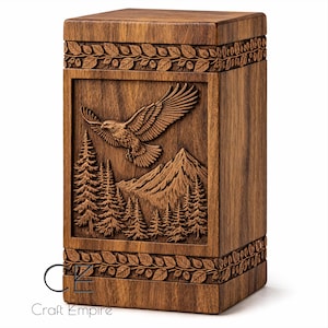 May include: A wooden box featuring a carved relief of an eagle soaring above mountains and pine trees. The rectangular box has leaf patterns at the top and bottom. The wood has a warm, natural colour.