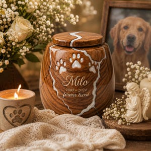 Könnte beinhalten: Eine Tierurne aus Holz mit polierter Oberfläche, weißer Harzeinlage, drei Pfotenabdrücken und der Gravur Milo 2008-2024 forever loved. Die Urne hat eine abgerundete Form mit passendem Deckel und einer deutlich sichtbaren Holzmaserung.