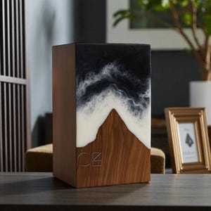 Puede incluir: Una caja rectangular de madera con un diseño de resina. La sección superior presenta un patrón de olas en blanco y negro, que se transforma en una forma de montaña en blanco, sobre una base de madera. Las letras "CZ" están grabadas en el frente.