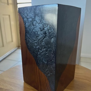 以下が含まれることがあります： 暗く質感のある木炭色のセンターパネルを備えた長方形の木箱。側面は光沢のある仕上げの濃い茶色です。木箱は木製のテーブルの上に置かれ、素材と質感のコントラストを示しています。