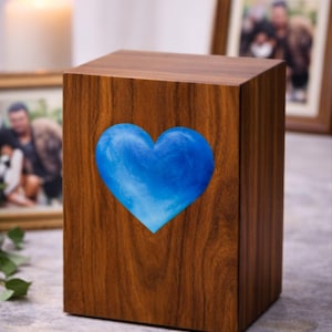 Pode incluir: Uma urna funerária de madeira com um design de coração azul. A urna retangular tem um acabamento de grão de madeira polida. Um coração azul está centrado na frente. Uma vela acesa e fotos emolduradas estão no fundo.
