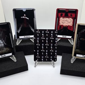 May include: Five playing cards displayed on clear stands, each with a unique design. The cards feature dark imagery and text labels like "Rock," "Scissors," "Bomb," and "Paper." A central card has a repeating pattern of white figures on a black background.