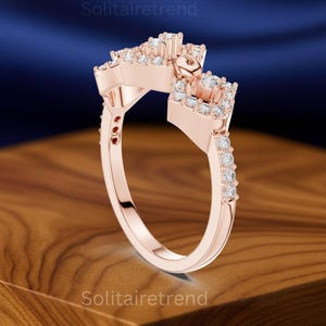 Puede incluir: Anillo de oro rosa con un dise&ntilde;o &uacute;nico, con m&uacute;ltiples piedras preciosas transparentes. El anillo tiene un engaste cuadrado y una banda adornada con piedras m&aacute;s peque&ntilde;as. El anillo se muestra sobre una superficie de madera con un fondo azul oscuro.