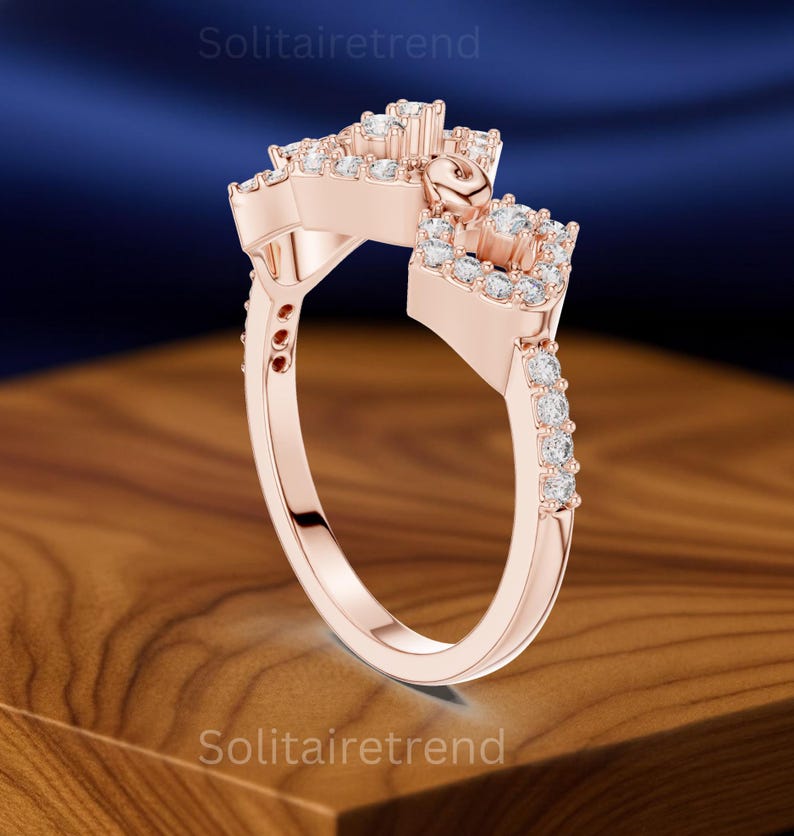 Puede incluir: Anillo de oro rosa con un dise&ntilde;o &uacute;nico, con m&uacute;ltiples piedras preciosas transparentes. El anillo tiene un engaste cuadrado y una banda adornada con piedras m&aacute;s peque&ntilde;as. El anillo se muestra sobre una superficie de madera con un fondo azul oscuro.