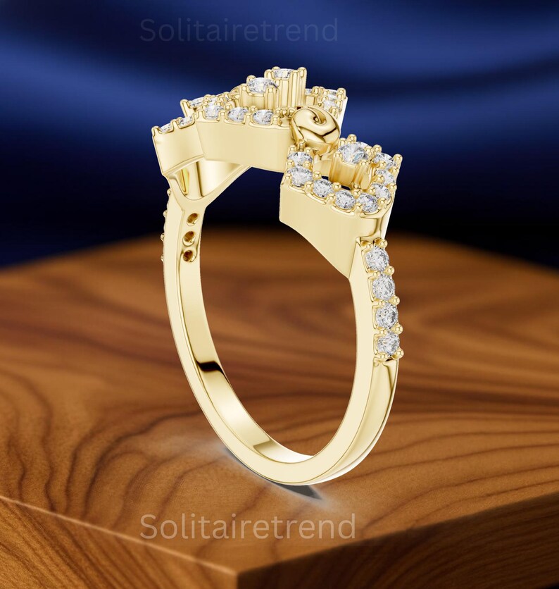 Puede incluir: Un anillo dorado con un dise&ntilde;o &uacute;nico, con m&uacute;ltiples piedras preciosas transparentes. El anillo tiene una ca&ntilde;a dividida y una parte superior decorativa con un elemento en forma de coraz&oacute;n. El anillo est&aacute; sobre una superficie de madera.