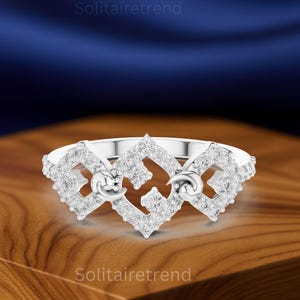 Puede incluir: Un anillo de plata con un dise&ntilde;o geom&eacute;trico, incrustado con numerosas piedras preciosas peque&ntilde;as y transparentes. El anillo presenta un dise&ntilde;o central con dos formas de media luna. El anillo se muestra sobre una superficie de madera.