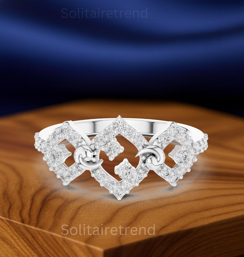 Puede incluir: Un anillo de plata con un dise&ntilde;o geom&eacute;trico, incrustado con numerosas piedras preciosas peque&ntilde;as y transparentes. El anillo presenta un dise&ntilde;o central con dos formas de media luna. El anillo se muestra sobre una superficie de madera.